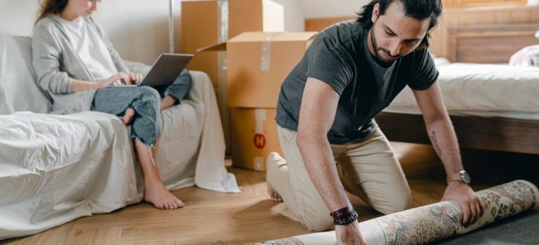 Man rolling up the carpet while his girlfriend is browsing the internet