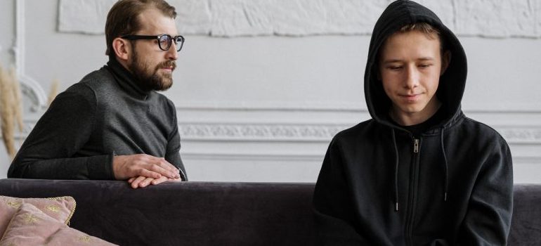 male in hoodie sitting on a sofa and a man with glasses behind